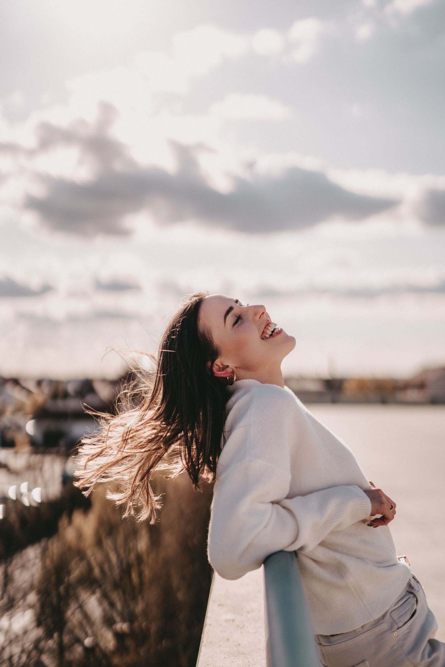 Joyful portrait leaning back against railing with hair blowing in wind at sunset.