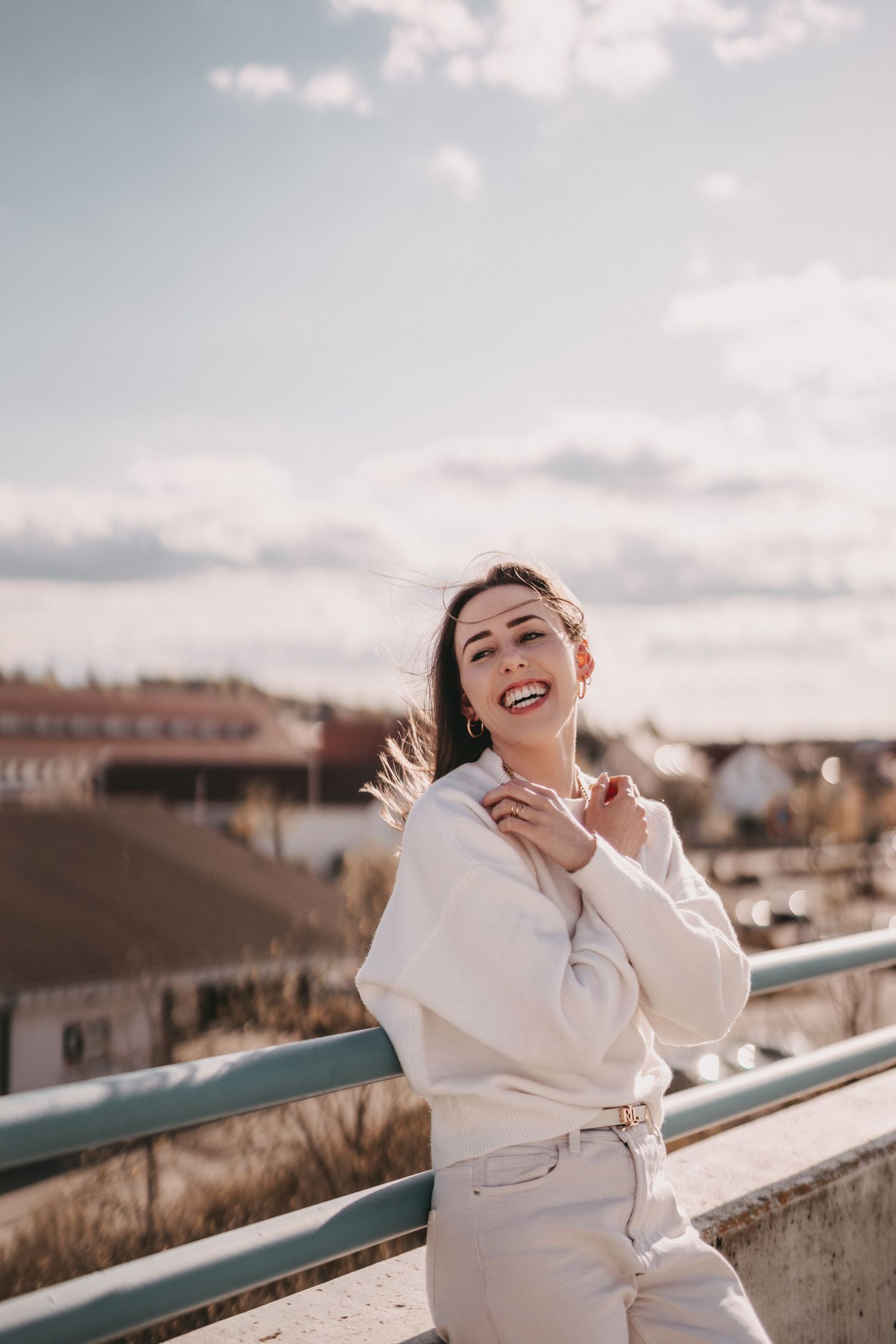 Candid laughing portrait in white sweater against cloudy sky on rooftop.