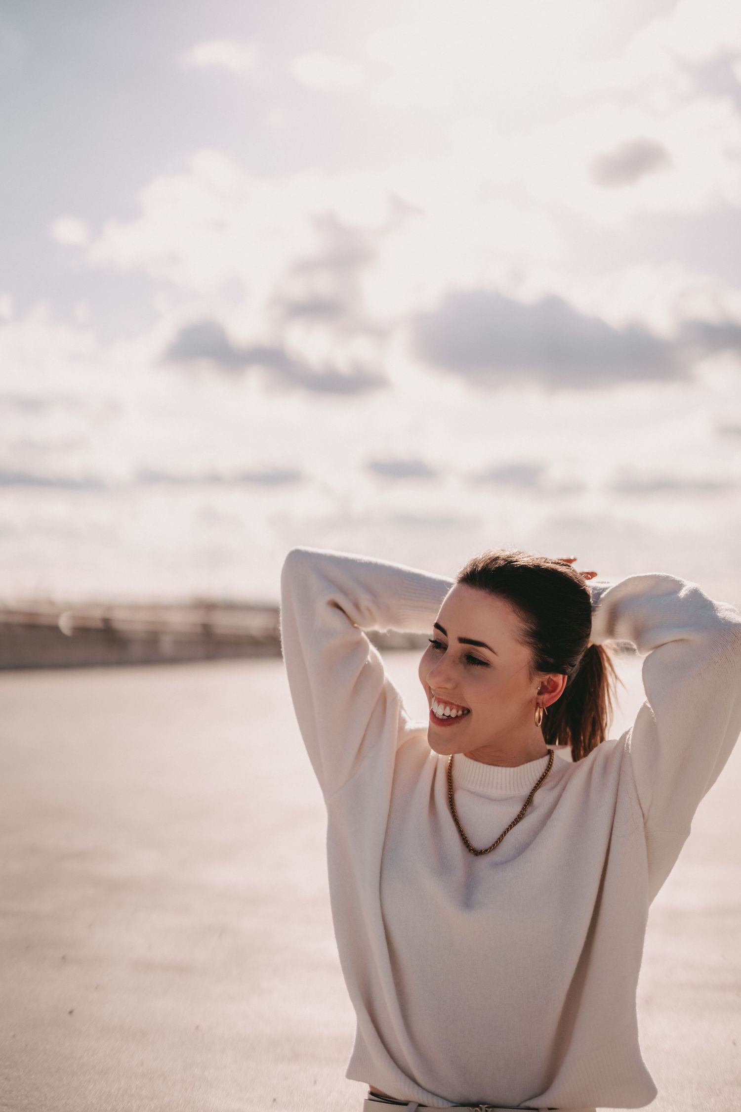 Carefree portrait in oversized white sweater with hands raised during golden hour.