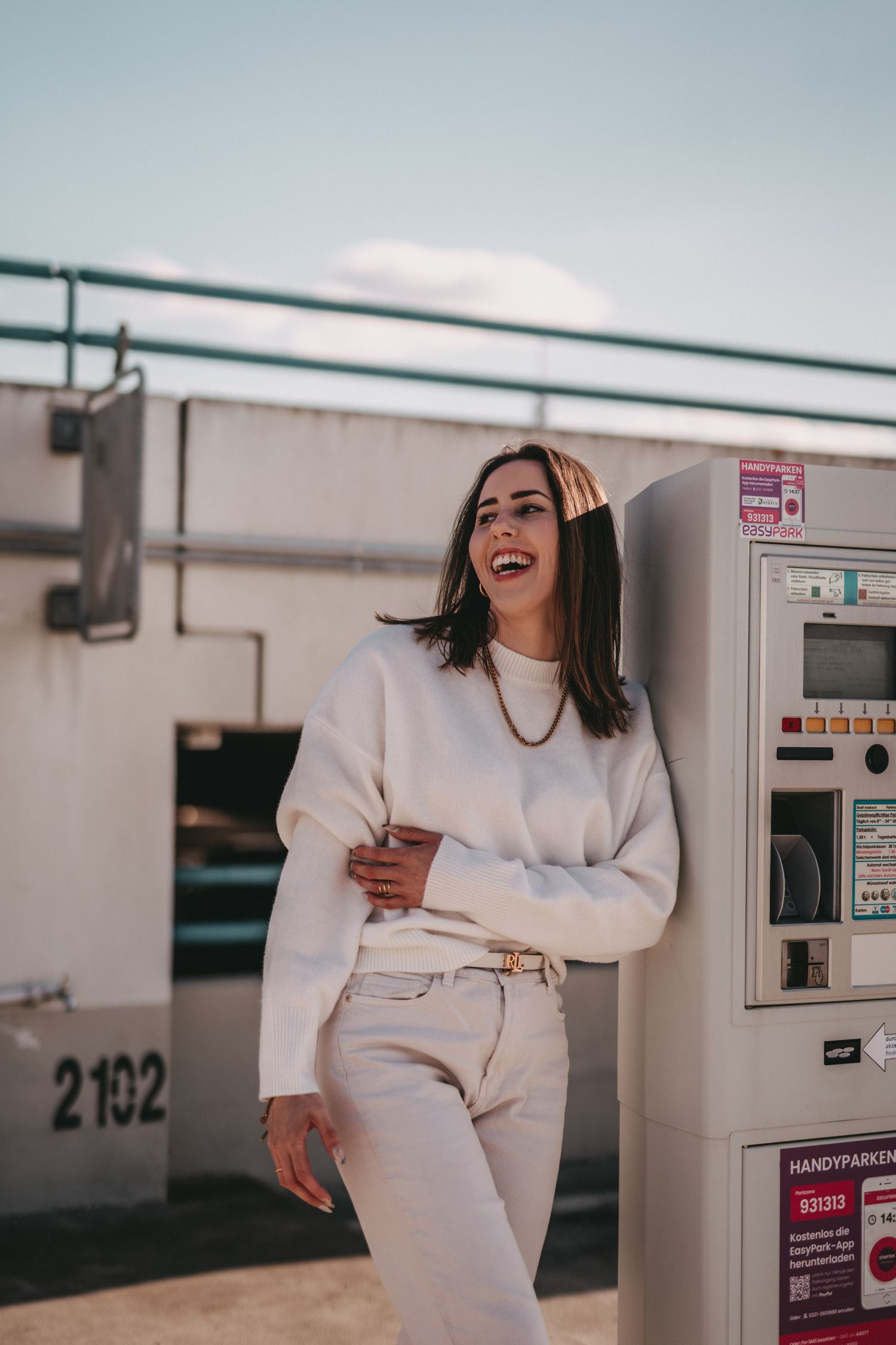Casual portrait in all-white outfit leaning against parking garage wall.