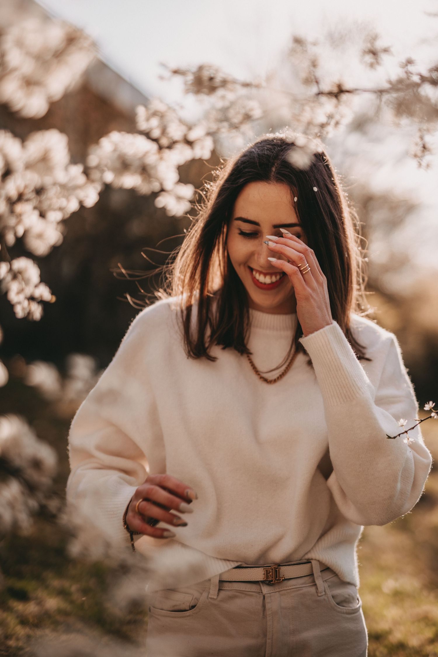 Candid laughing portrait in white sweater with cherry blossoms in background.