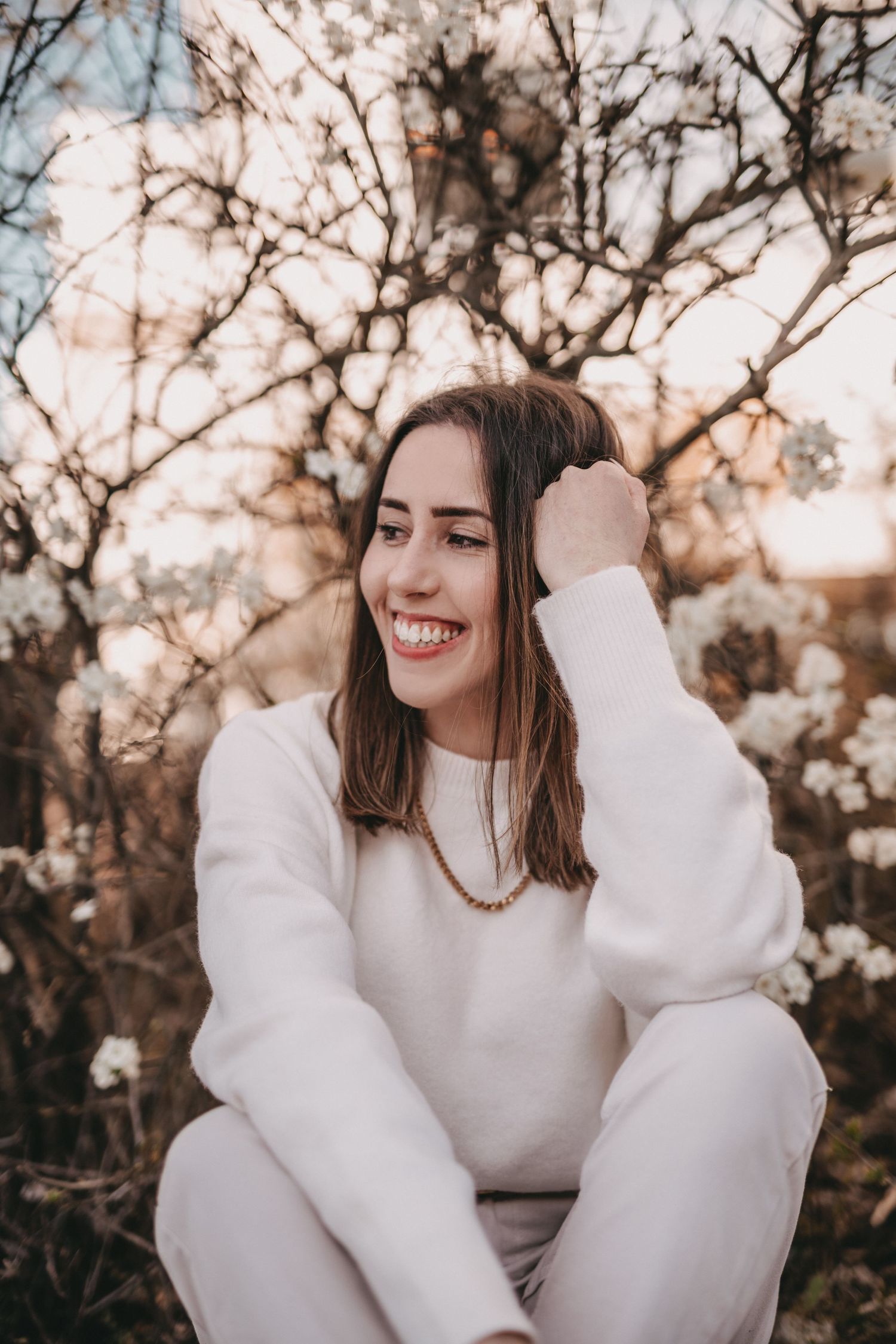 Natural portrait in white sweater sitting among blooming spring branches at sunset.
