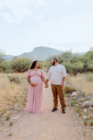 A couple walks hand in hand through a desert trail with mountains and cacti in the distance.