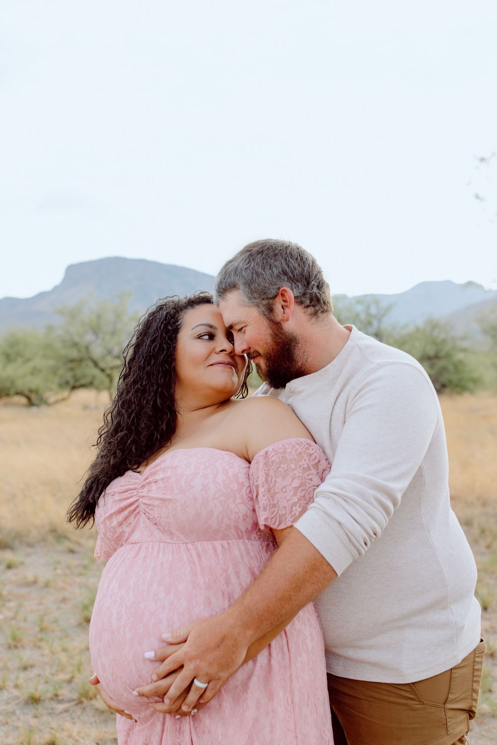Romantic couple embracing during desert maternity photoshoot in pink dress.