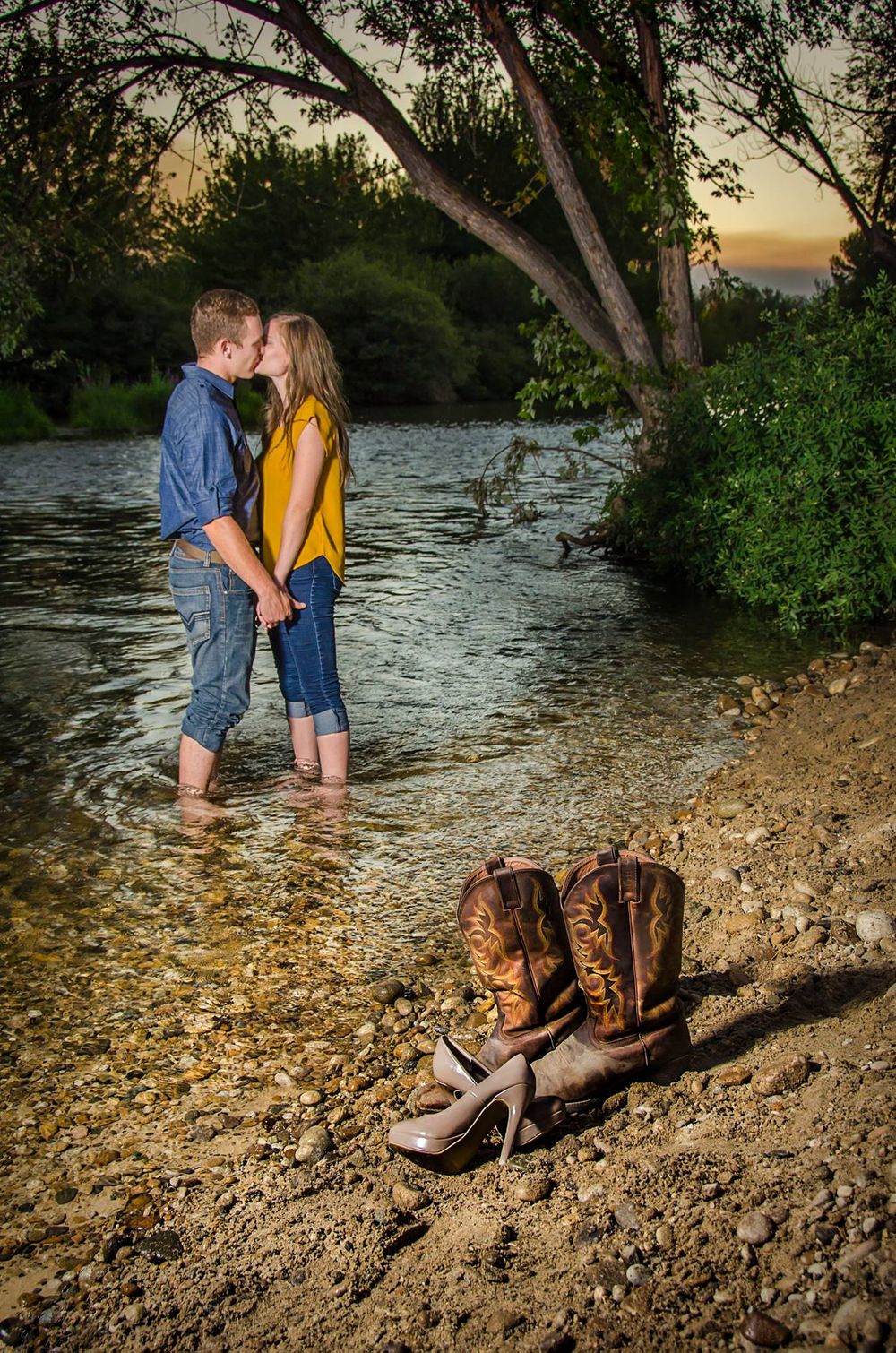 Romantic couple standing in creek water at sunset with boots and shoes on shore.
