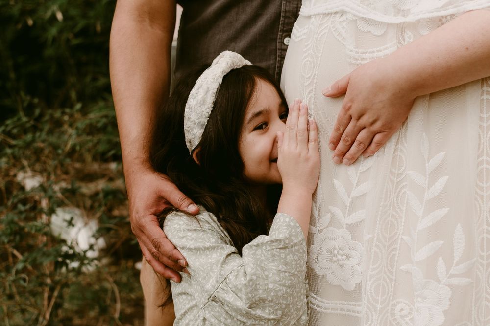 A young child kisses a pregnant belly while standing with parents.