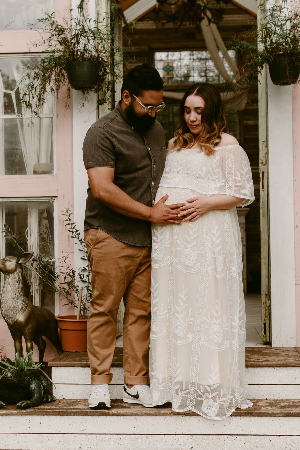 A couple shares a tender moment during their maternity photoshoot on a charming porch with hanging plants.