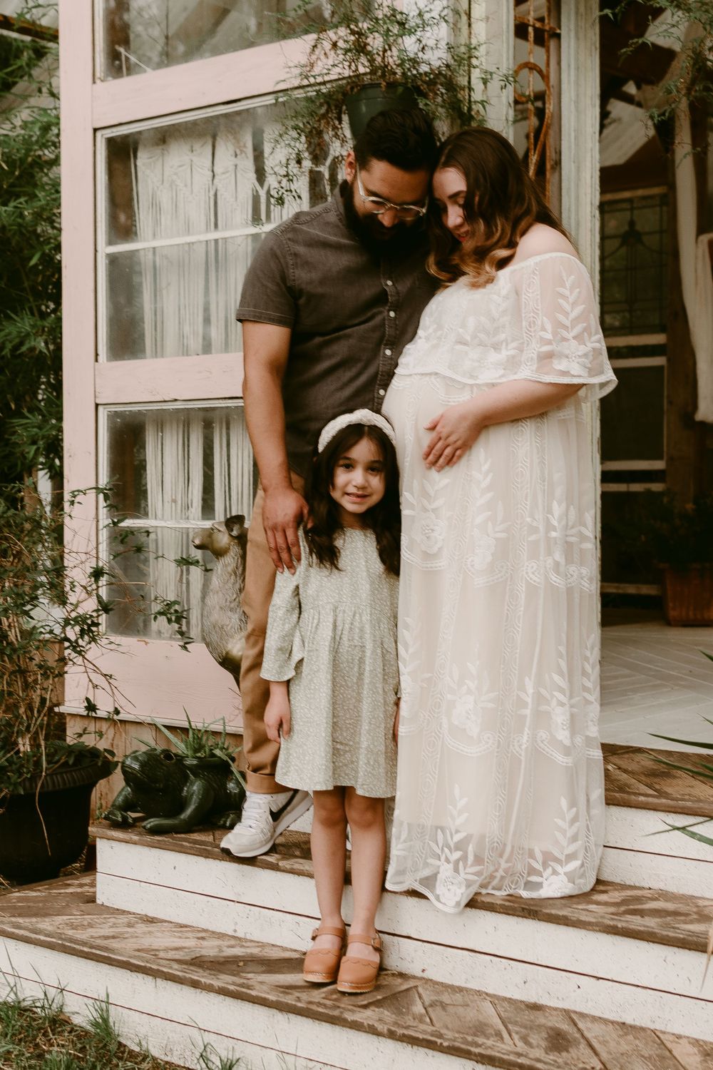 A loving family poses together on their front porch steps with a beautiful white lace maternity dress.