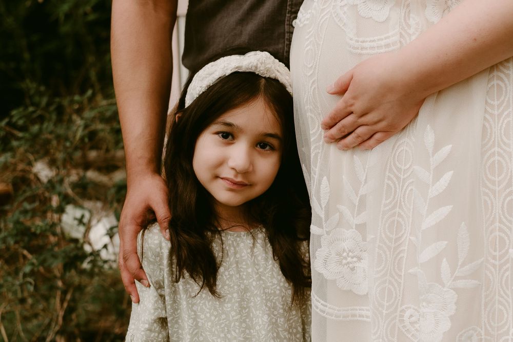 A little girl in a white dress stands between two adults.