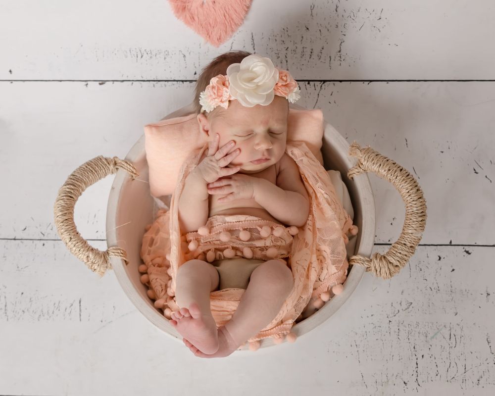 Newborn baby wearing peach outfit rests in woven basket on white wooden floor with floral headband.