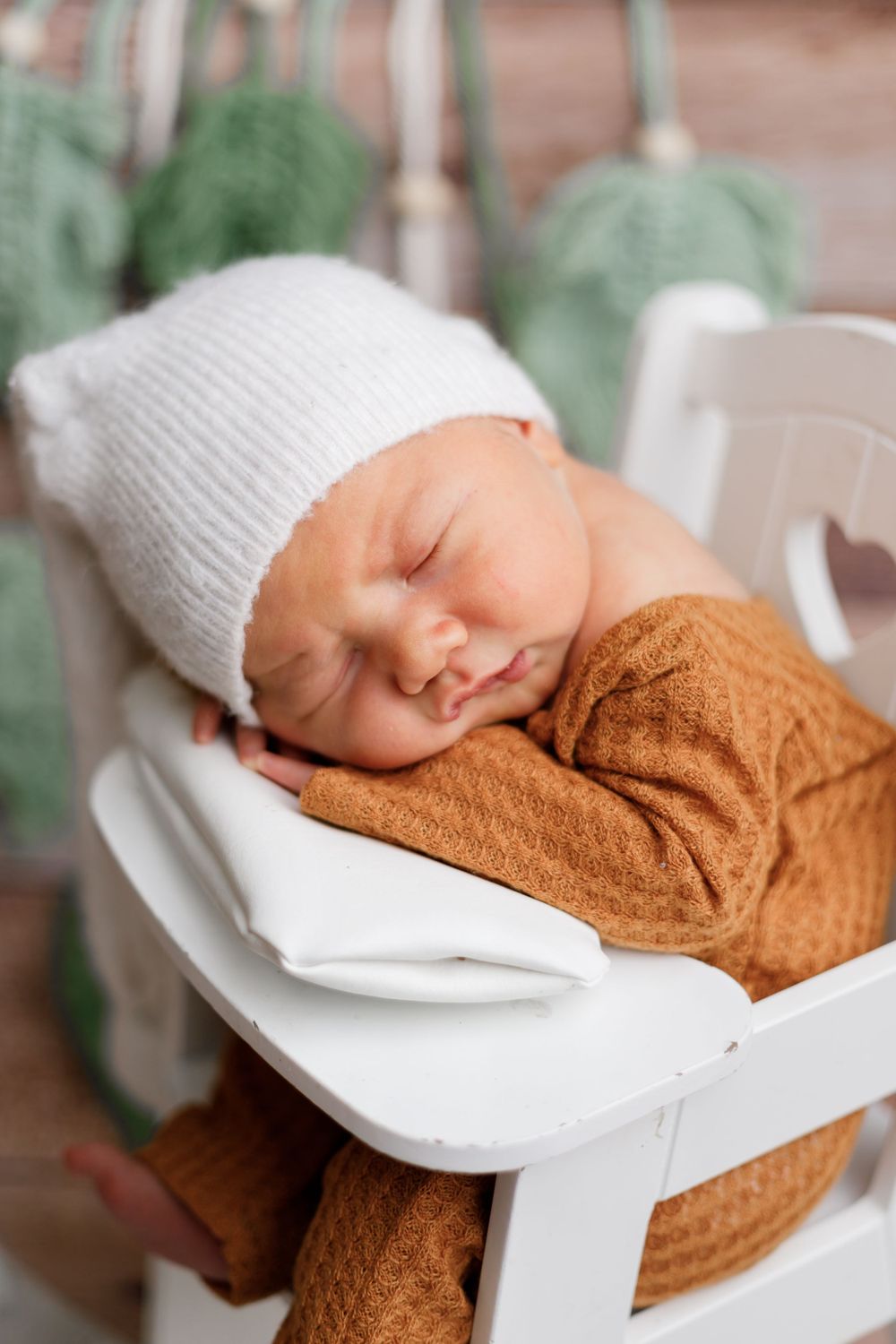 Baby sleeping in white chair wearing knit hat and brown outfit with green plants in background.