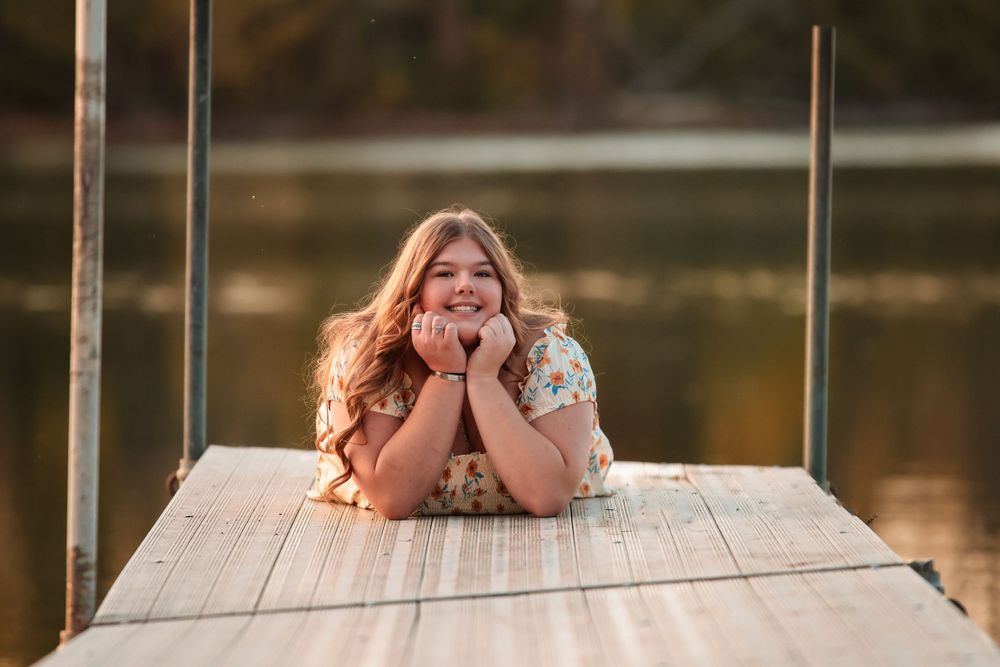 Person laying on a wooden dock smiling with autumn trees reflected in the calm lake water below.
