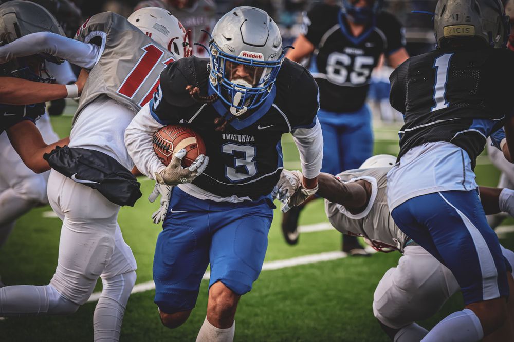 Football player in blue jersey carrying ball through defensive players during game action.