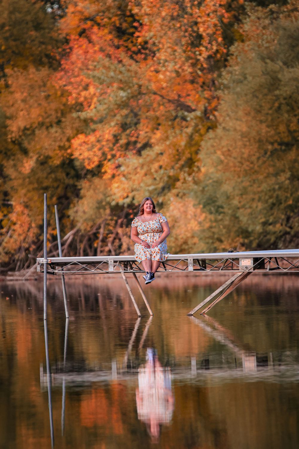 A person in a floral dress poses on a wooden dock over a lake during autumn with orange foliage reflecting in the water.
