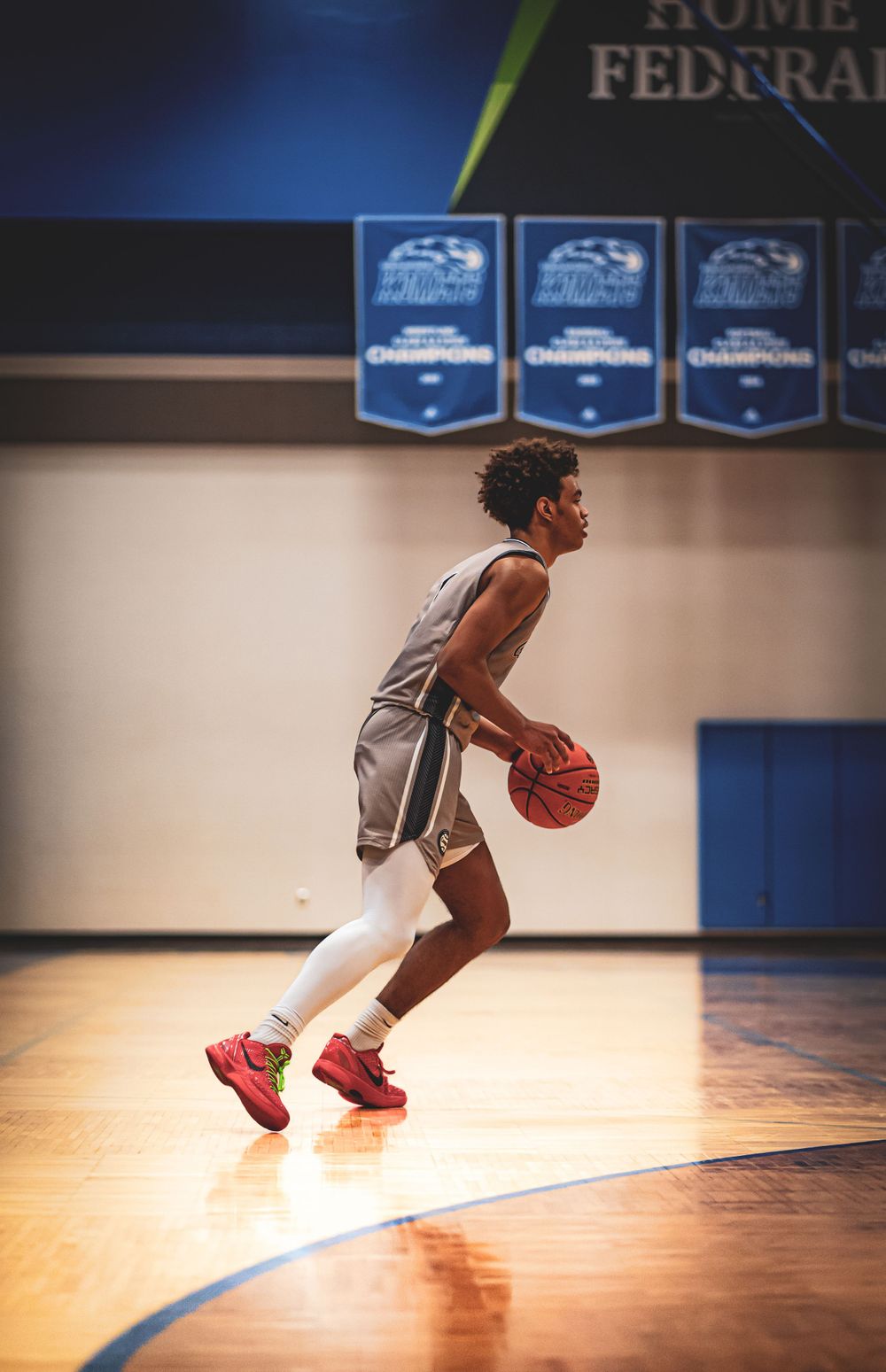 Basketball player dribbling on indoor court with championship banners overhead.