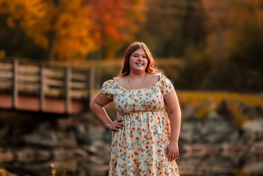 Person in floral dress poses against autumn scenery with bridge and colorful fall foliage.