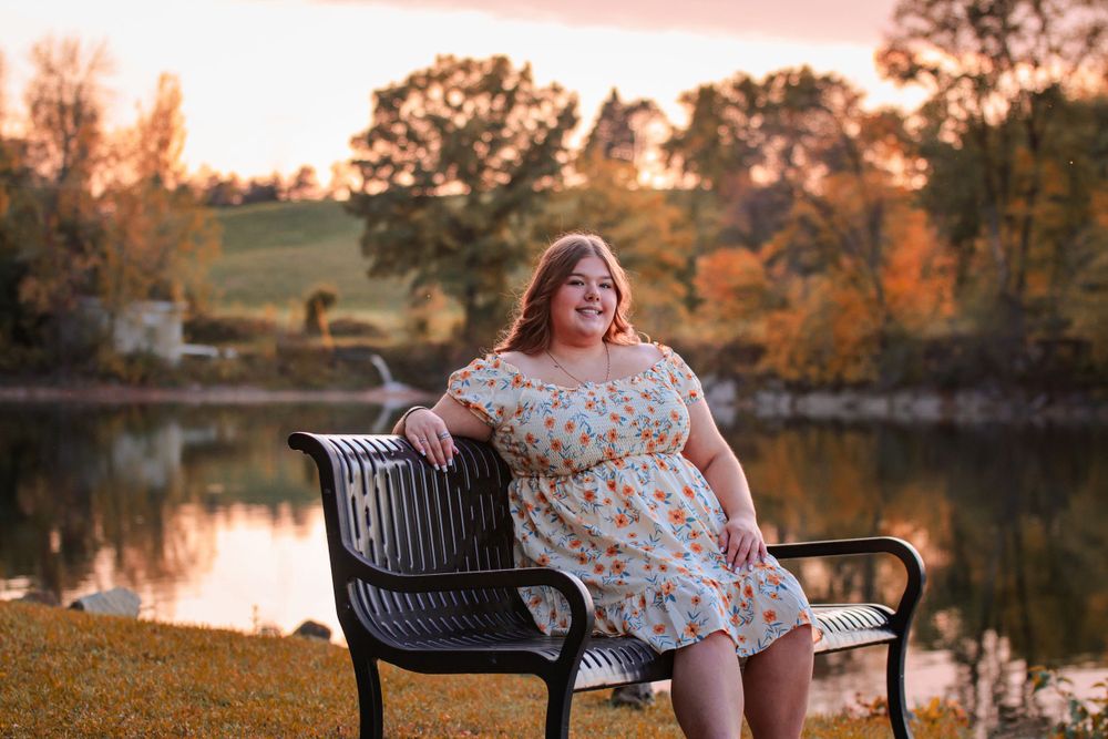 Person in floral dress sits on park bench by lake during golden hour with autumn colors in background.