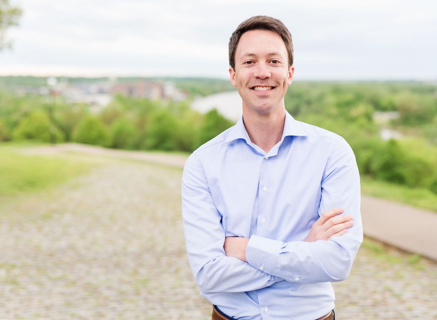 Professional portrait of person in light blue shirt standing outdoors with arms crossed.