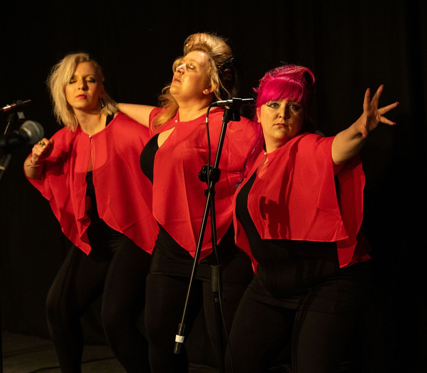 Three performers in matching red tops sing together on stage during a performance.