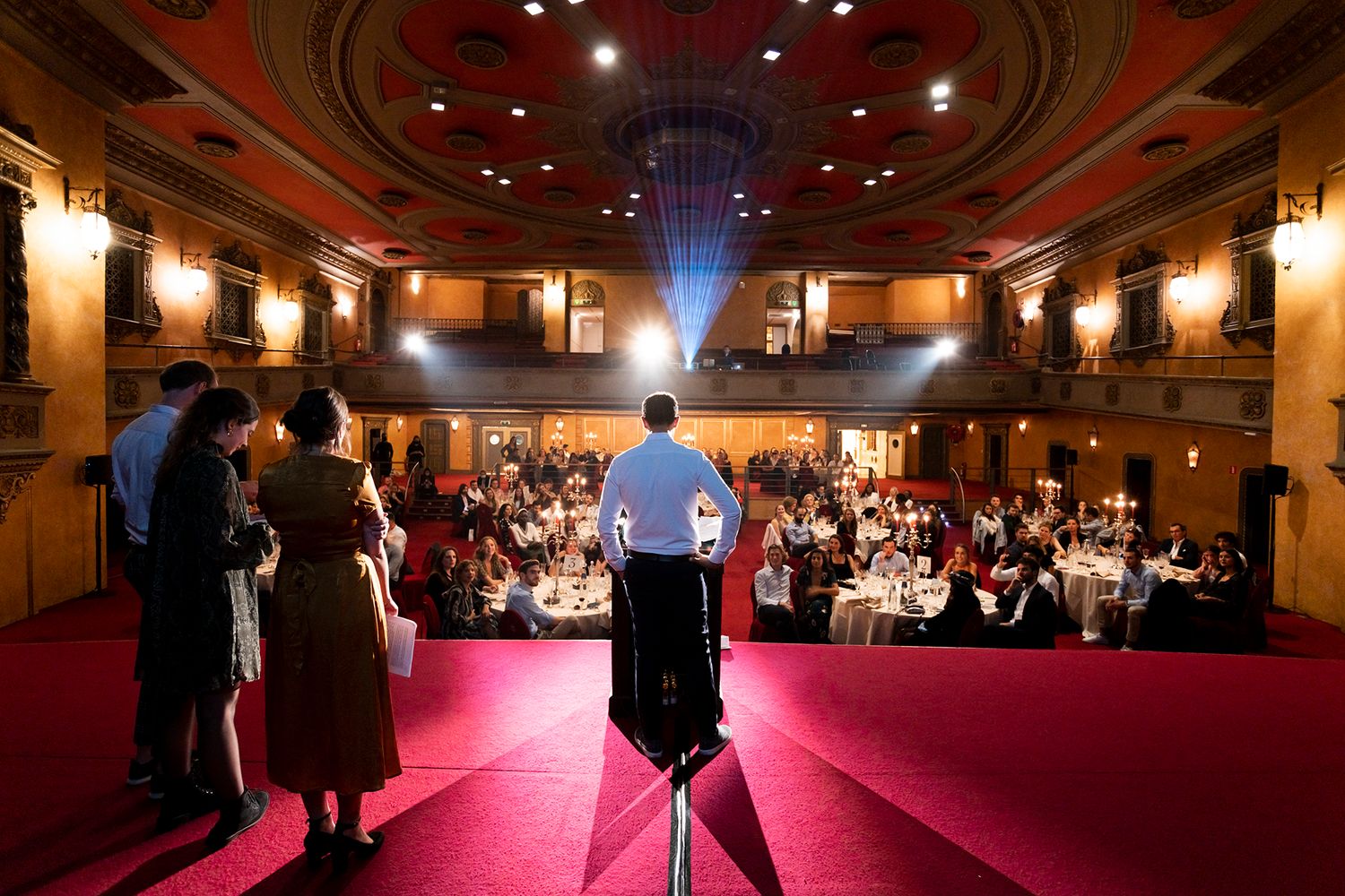 Speaker addresses audience from red-carpeted stage in ornate theater with balcony seating.