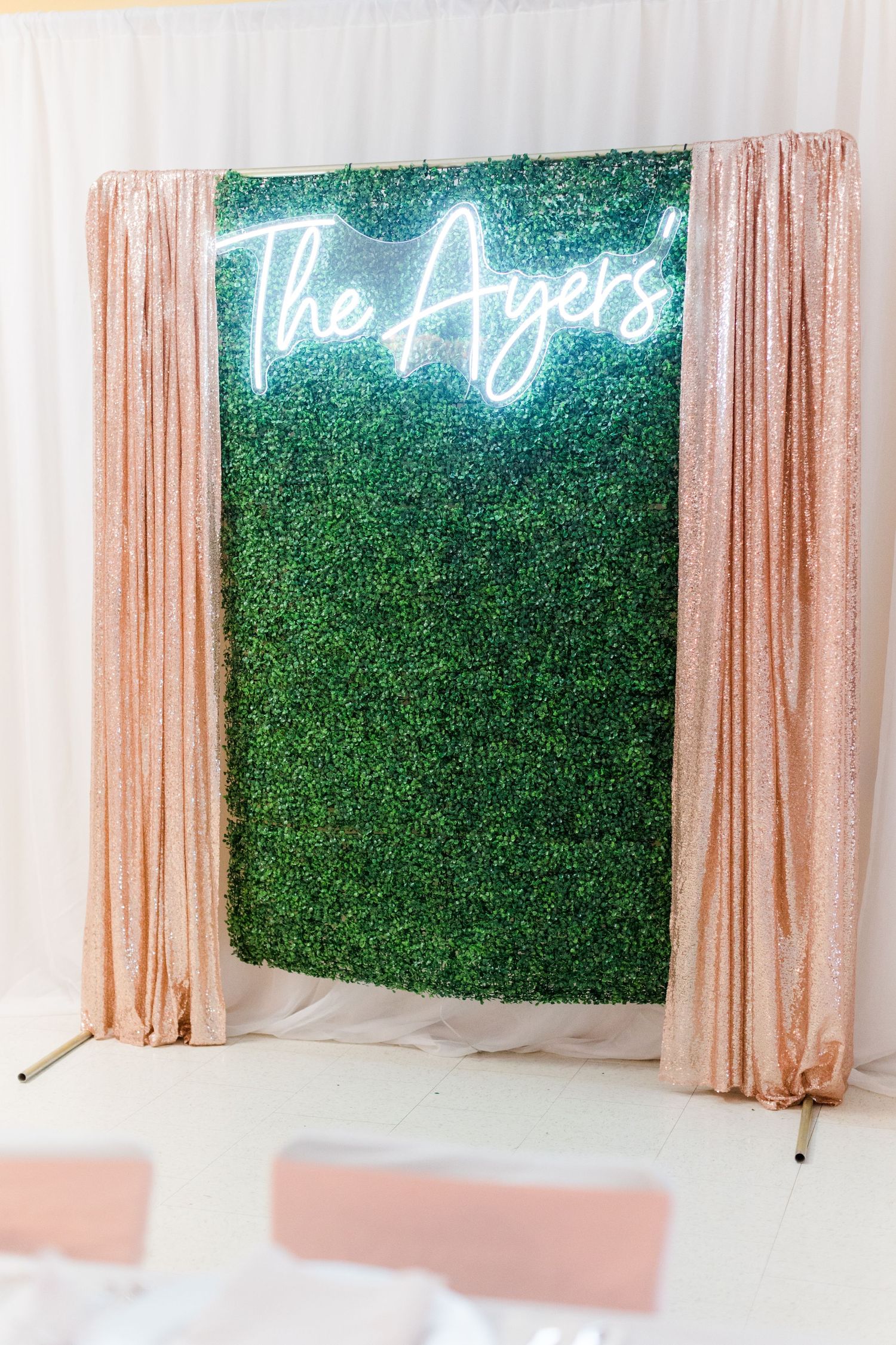 A green hedge wall backdrop with neon sign reading 'The Ayers' flanked by blush pink curtains.