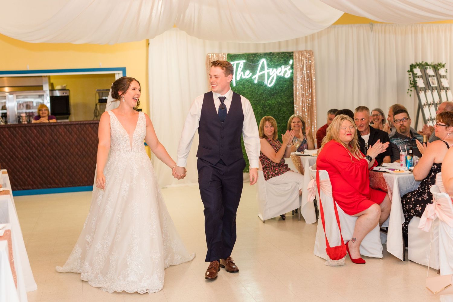 A wedding couple enters their reception venue decorated with a green hedge wall and neon sign.