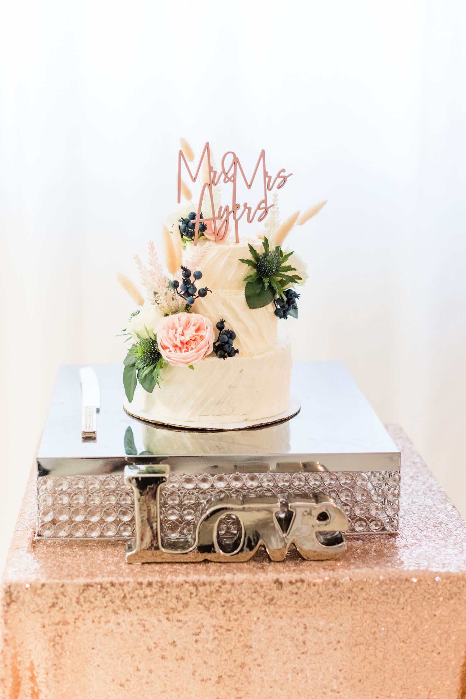 White wedding cake decorated with pink flowers and berries on a crystal love stand.