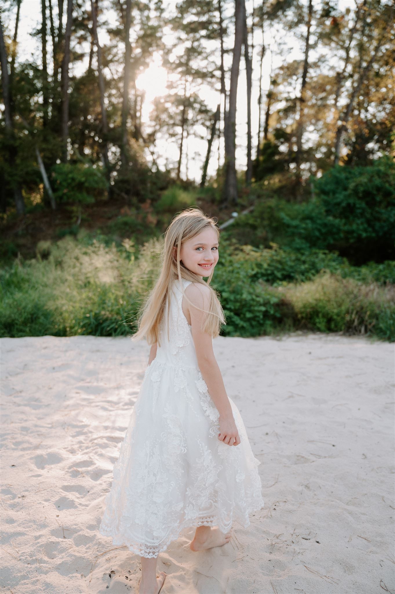 Young girl in white lace dress poses on sandy path with sunlit forest behind her.