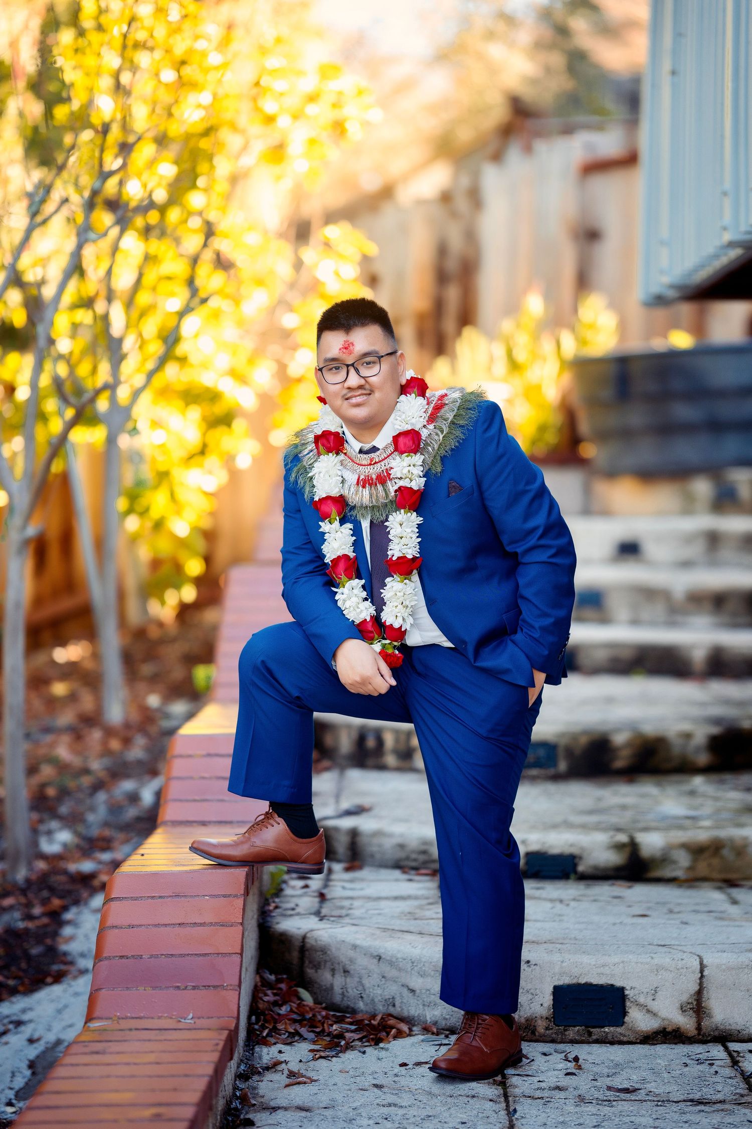A groom in a navy blue suit with traditional flower garlands poses on stone steps.