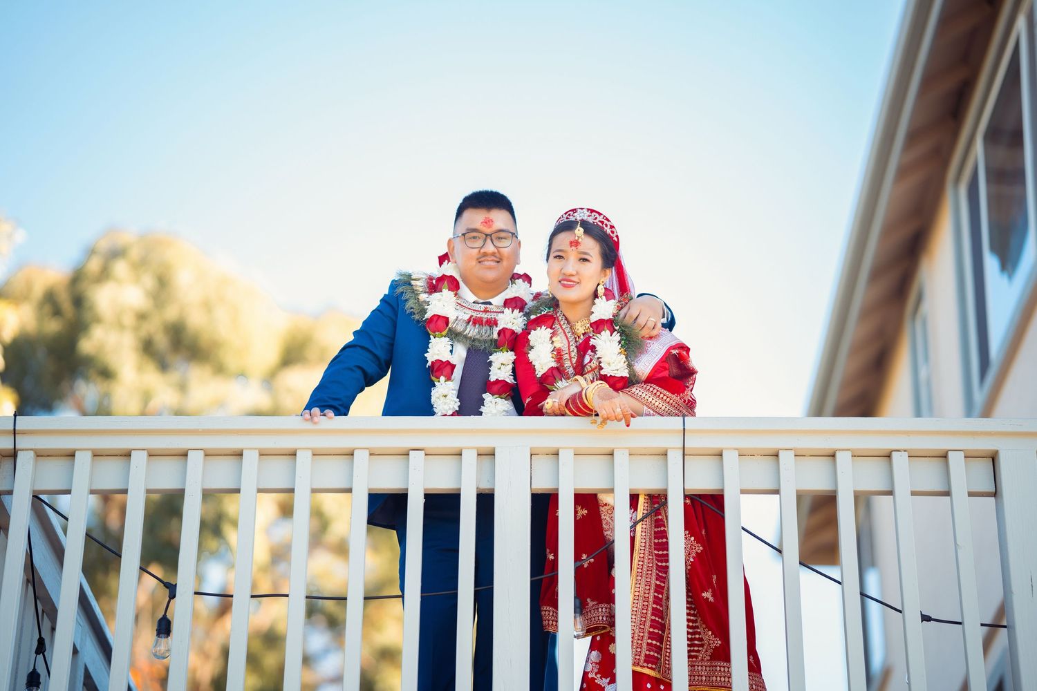 A couple in fusion wedding attire poses together on a white balcony.