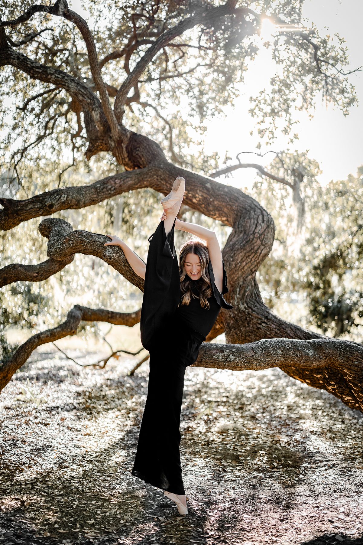 Dancer in black performs graceful pose against sunlit tree backdrop.