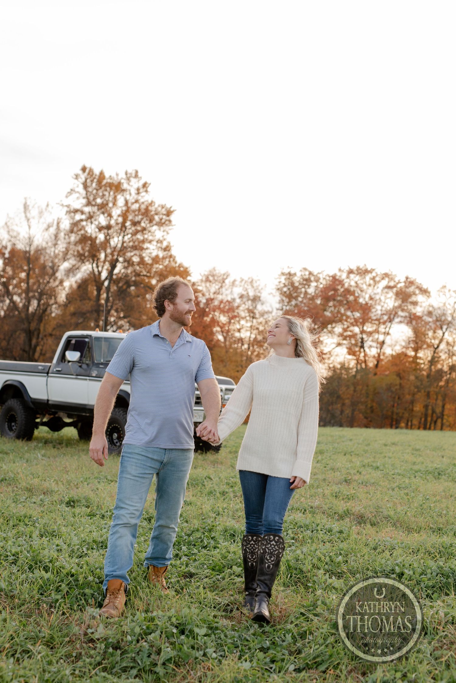 Couple walking hand in hand through autumn field with classic pickup truck in background.
