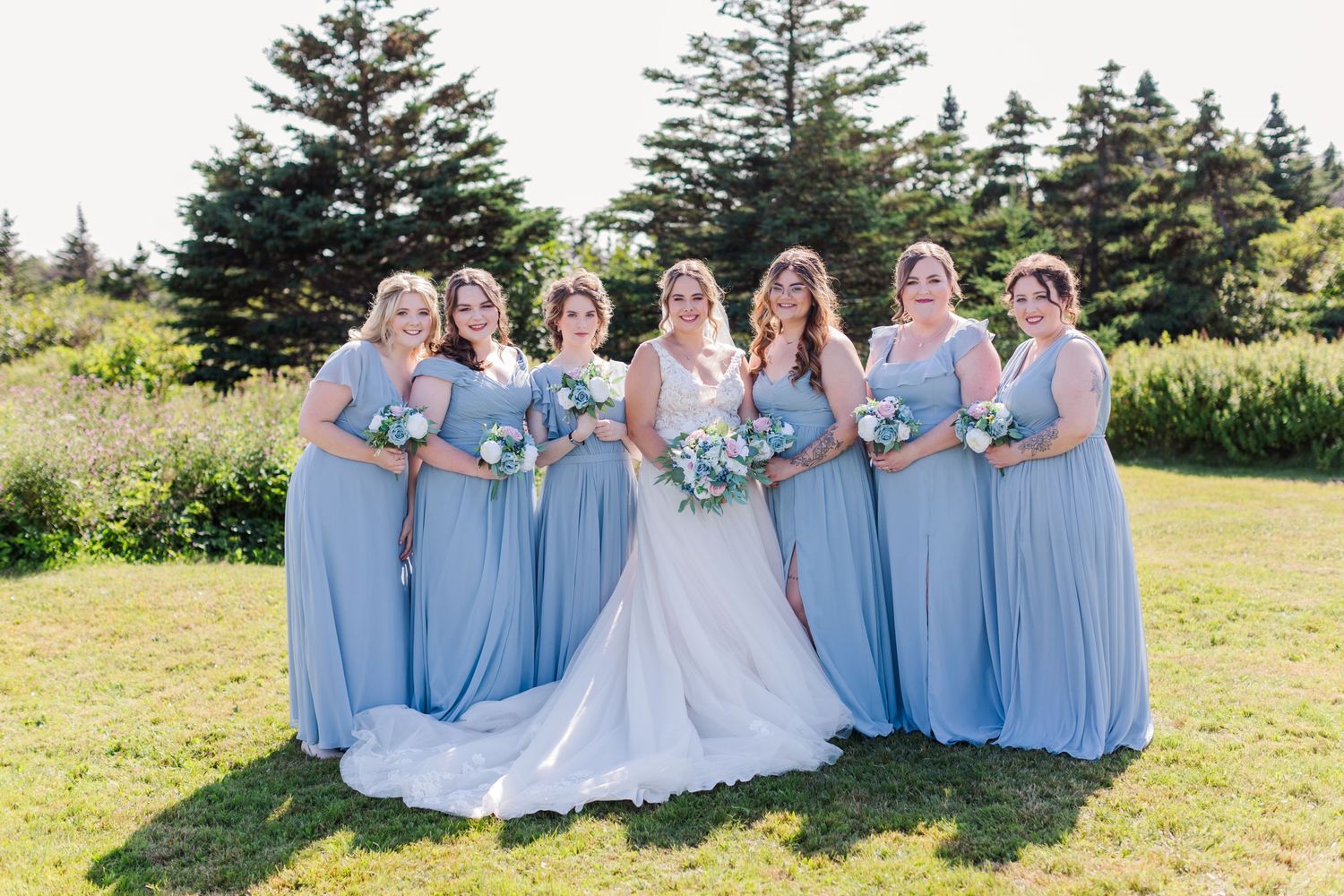 Bridesmaids in powder blue dresses pose with bride on sunny outdoor lawn.