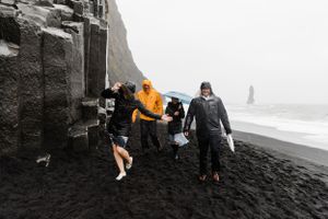 A group walks along a dramatic black sand beach with steep cliffs and sea stacks in the misty background.