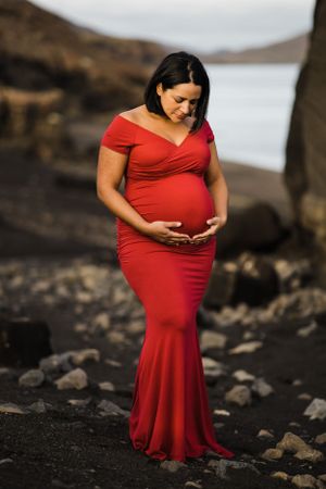 Expectant mother in red dress cradling baby bump against rocky coastal backdrop.