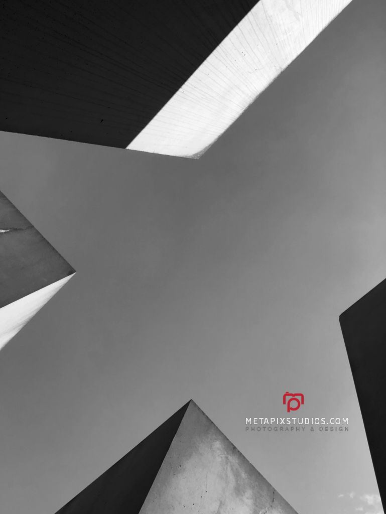 Black and white upward view of angular concrete structures against a bright sky at Berlin Holocaust Memorial.