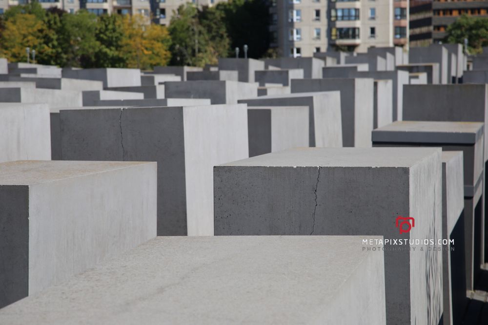 Color photo showing rows of gray concrete stelae with city buildings visible in the background in Berlin.