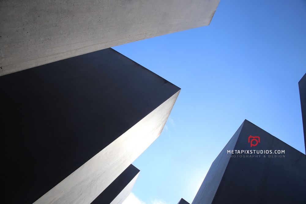 Upward view of dark angular concrete blocks against a bright blue sky at the Holocaust Memorial Berlin.