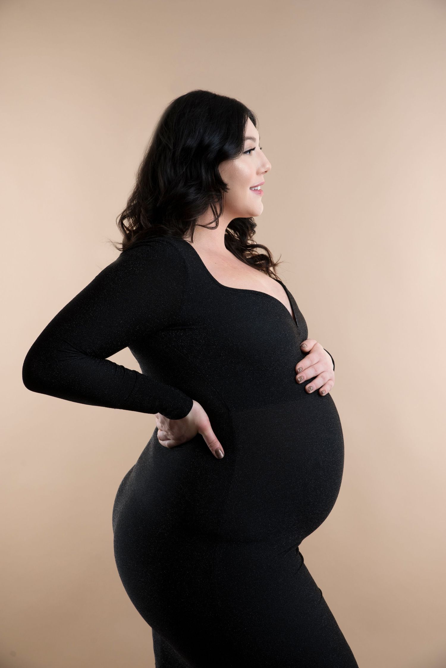 Side view maternity portrait of woman in black dress against neutral background.