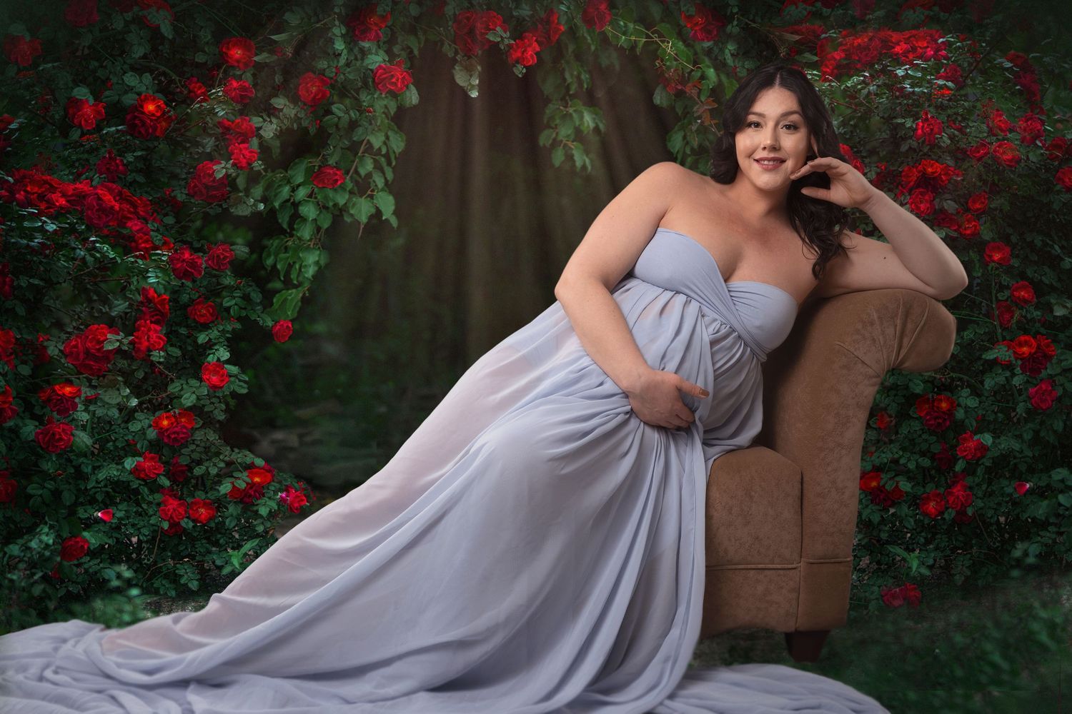 Maternity portrait of woman in grey gown reclining on chair among red roses.