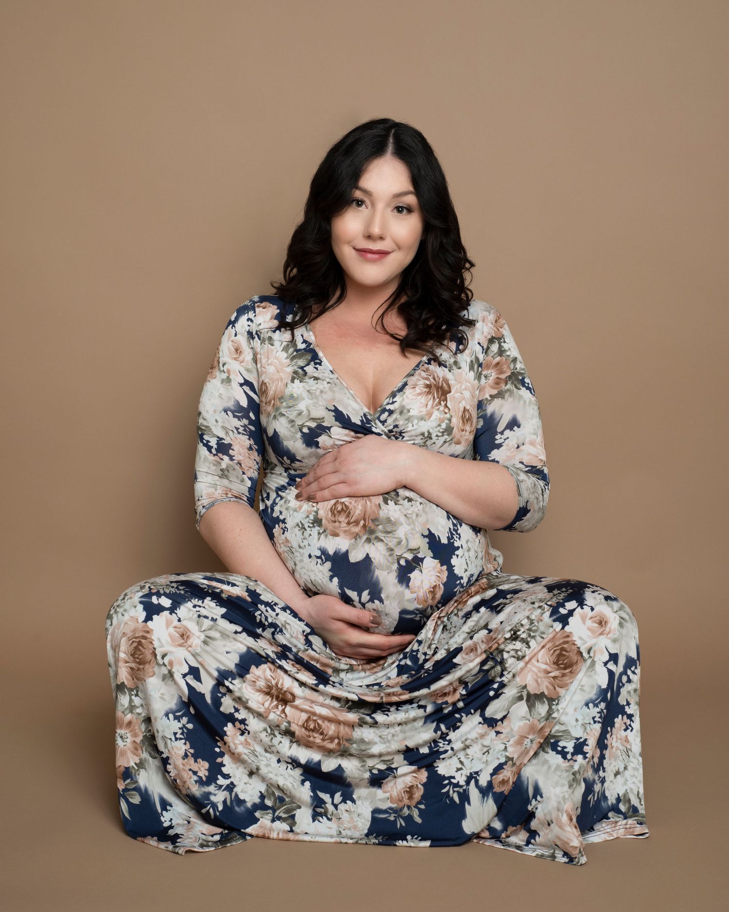 Maternity portrait of woman in floral dress sitting against beige background.