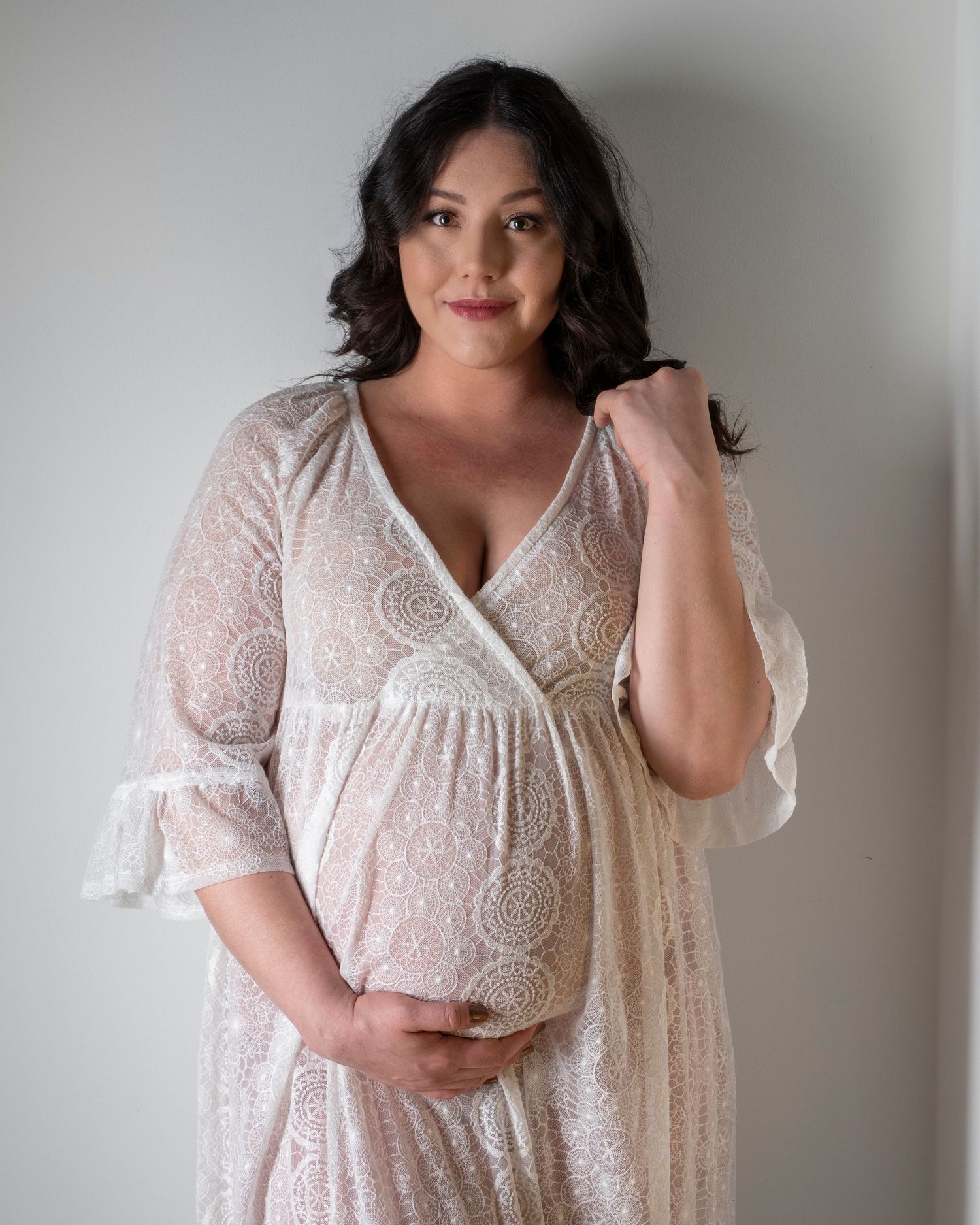 Maternity portrait of person in white lace dress cradling baby bump against light grey wall.