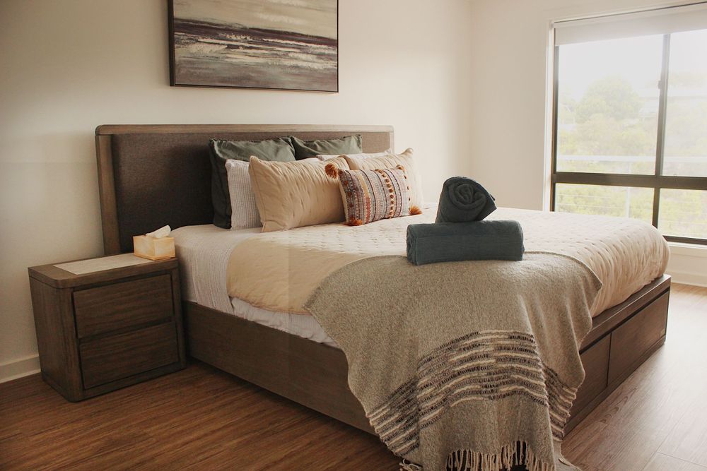 Modern bedroom interior with hardwood floors, white bedding, and large windows offering natural light.
