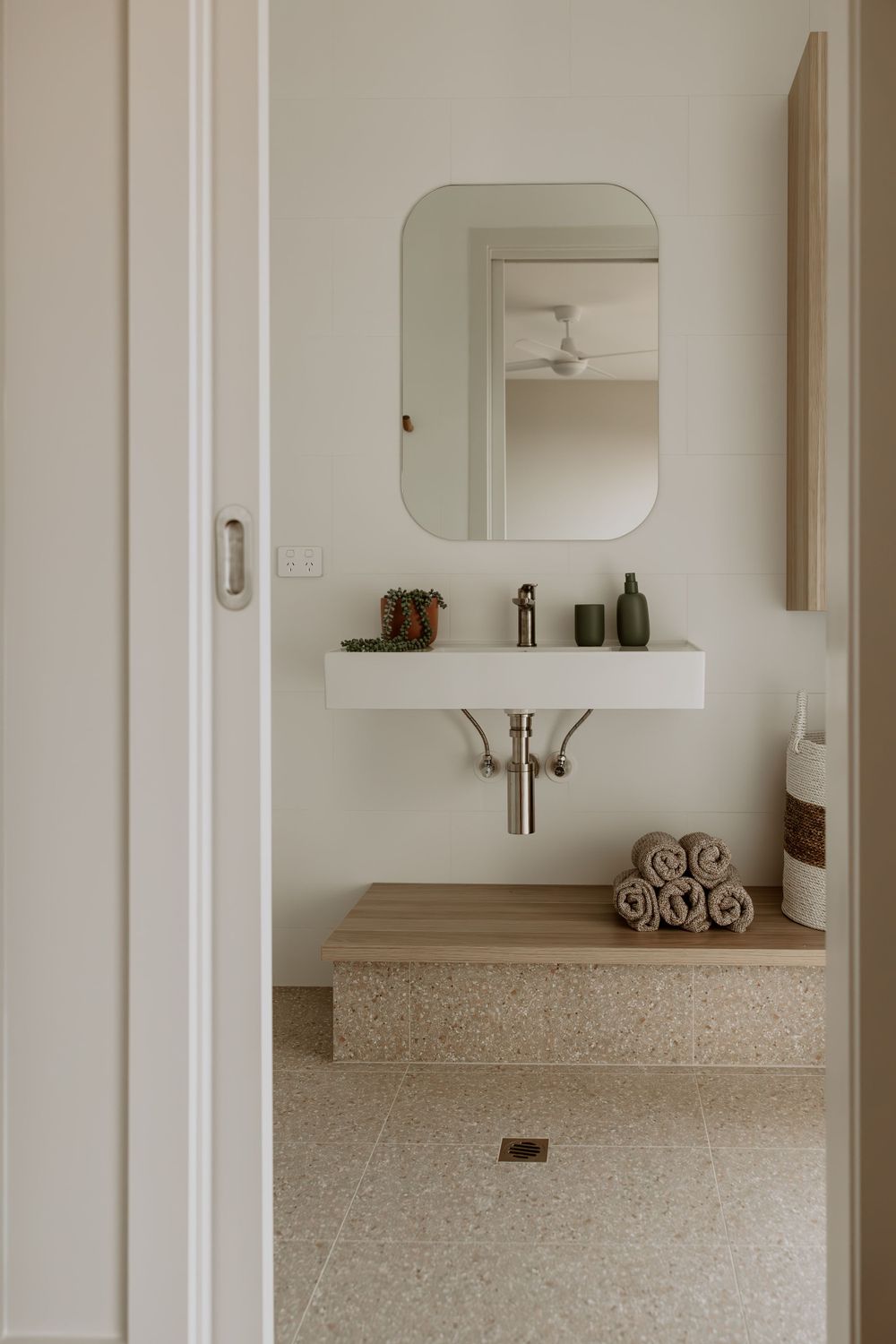 Modern bathroom interior with curved mirror, wooden shelving, and neutral color palette.