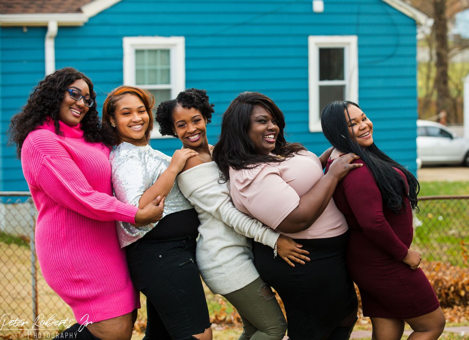 Five women laugh and embrace playfully outdoors near a blue house in colorful outfits.