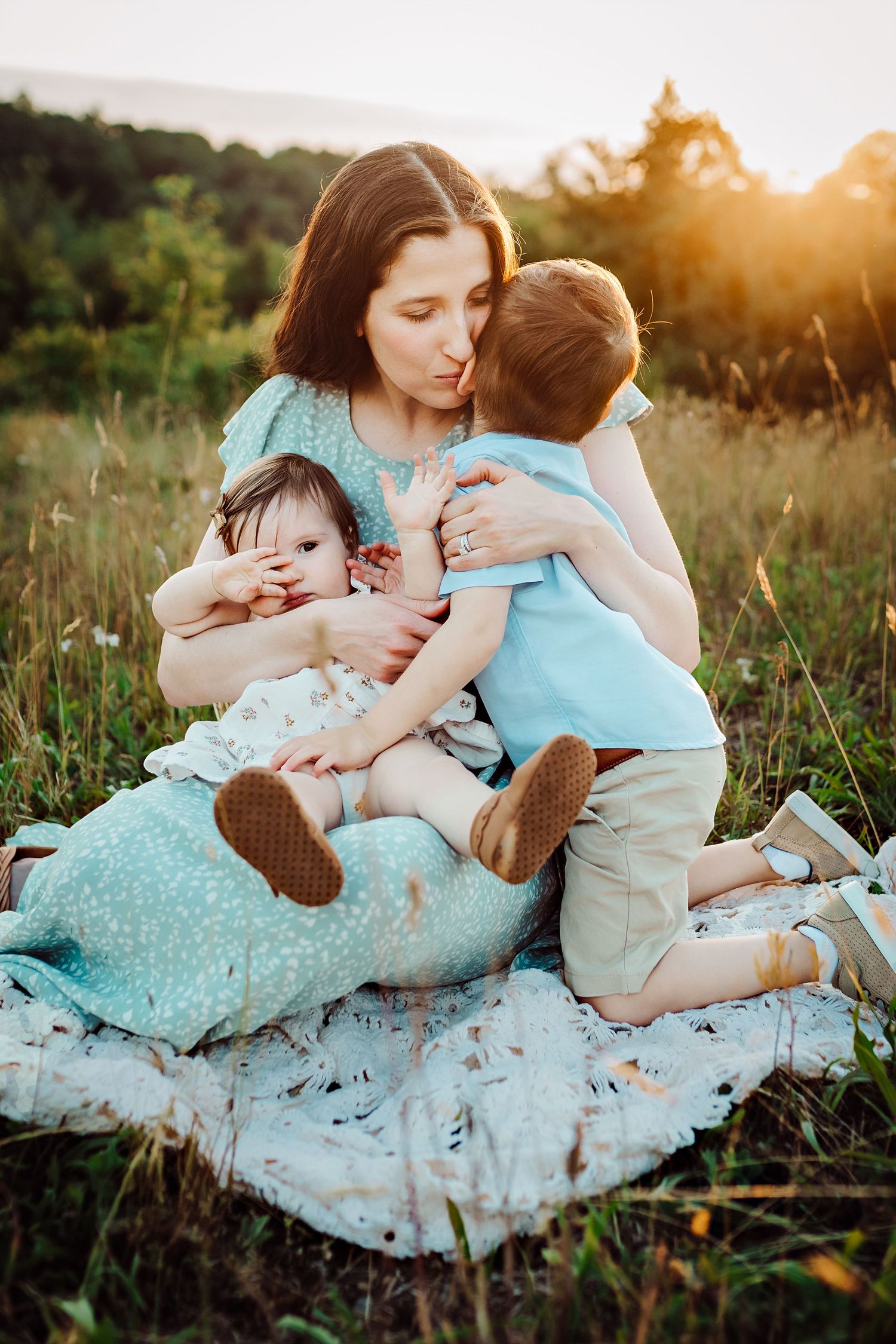 Family sharing tender moment while sitting on blanket in grassy field at sunset.