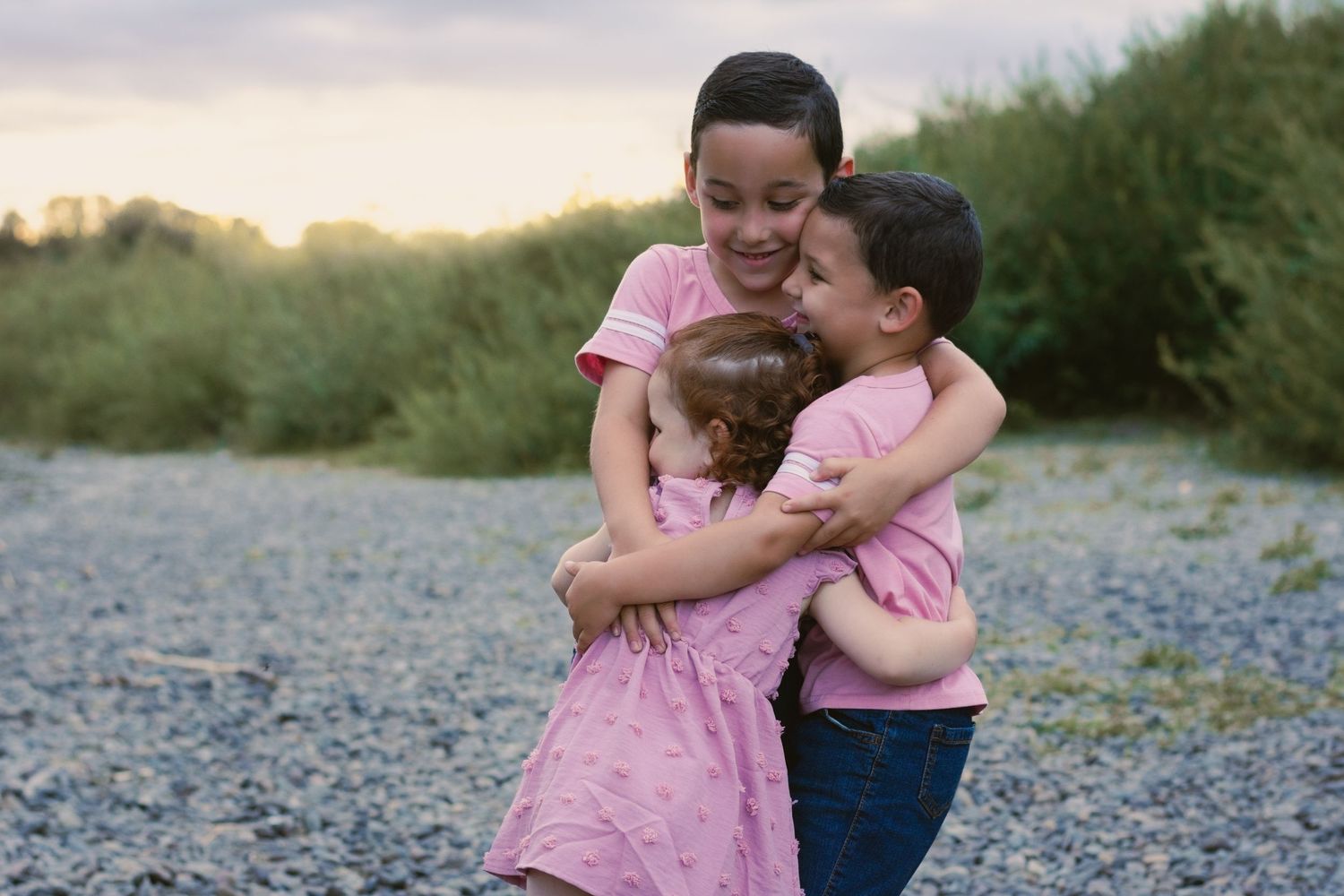 Siblings wearing matching outfits soft pink all hugging each other and smiling. by river on river bank. soft pink.