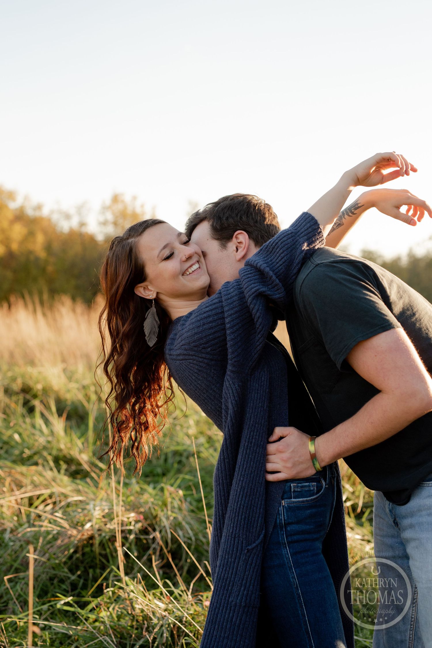 Romantic couple embraces in a golden field at sunset wearing casual clothes and a navy cardigan.
