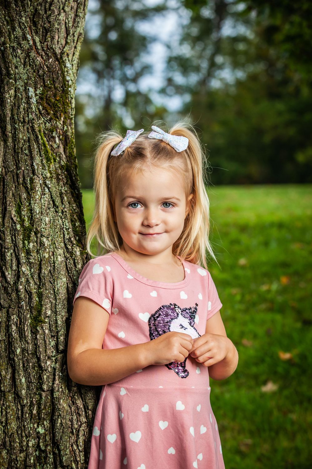Outdoor portraits near a tree trunk showing a pink heart-patterned dress and white hair bows.