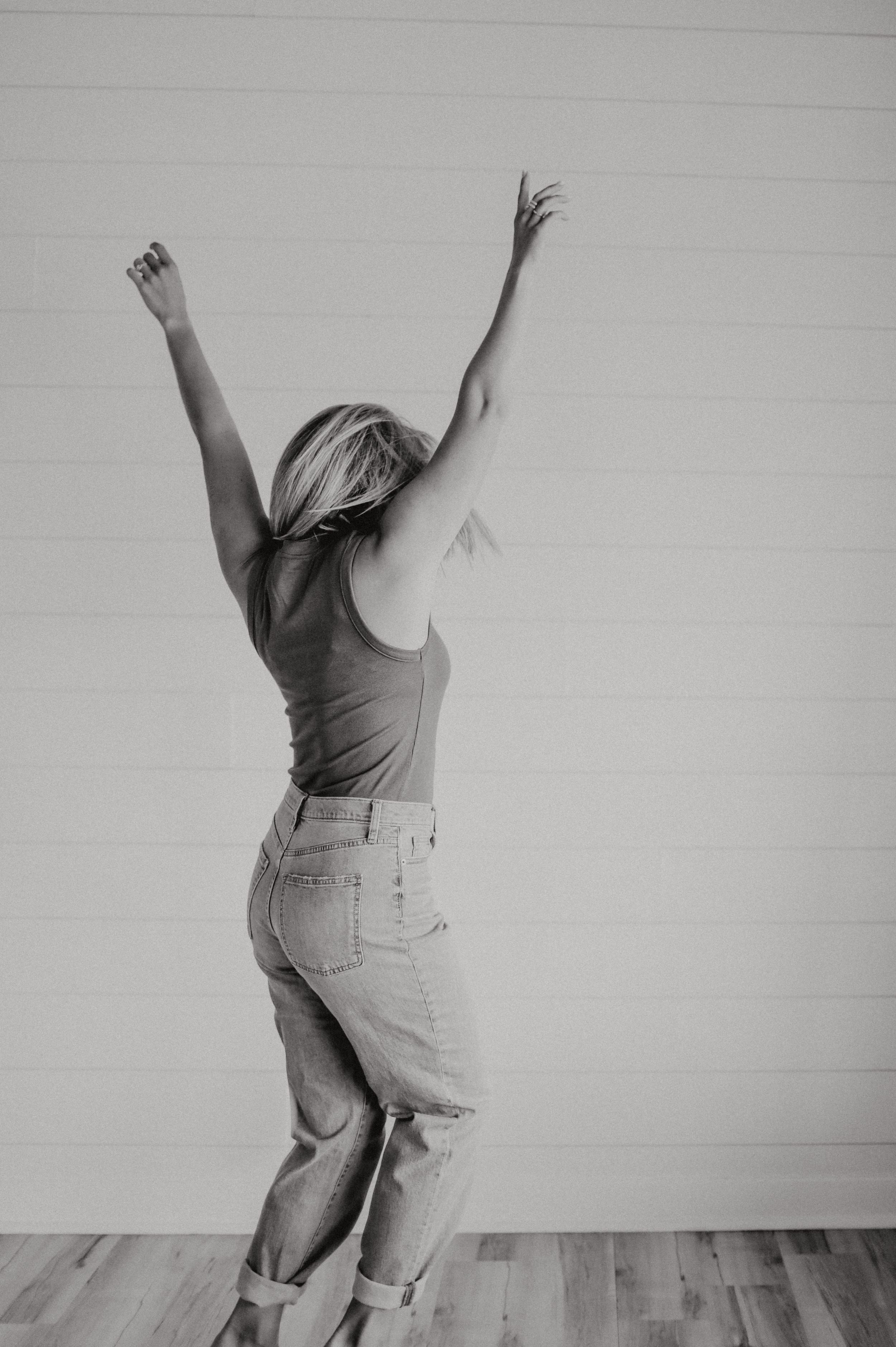 Black and white photo of person with raised arms dancing against white wall.